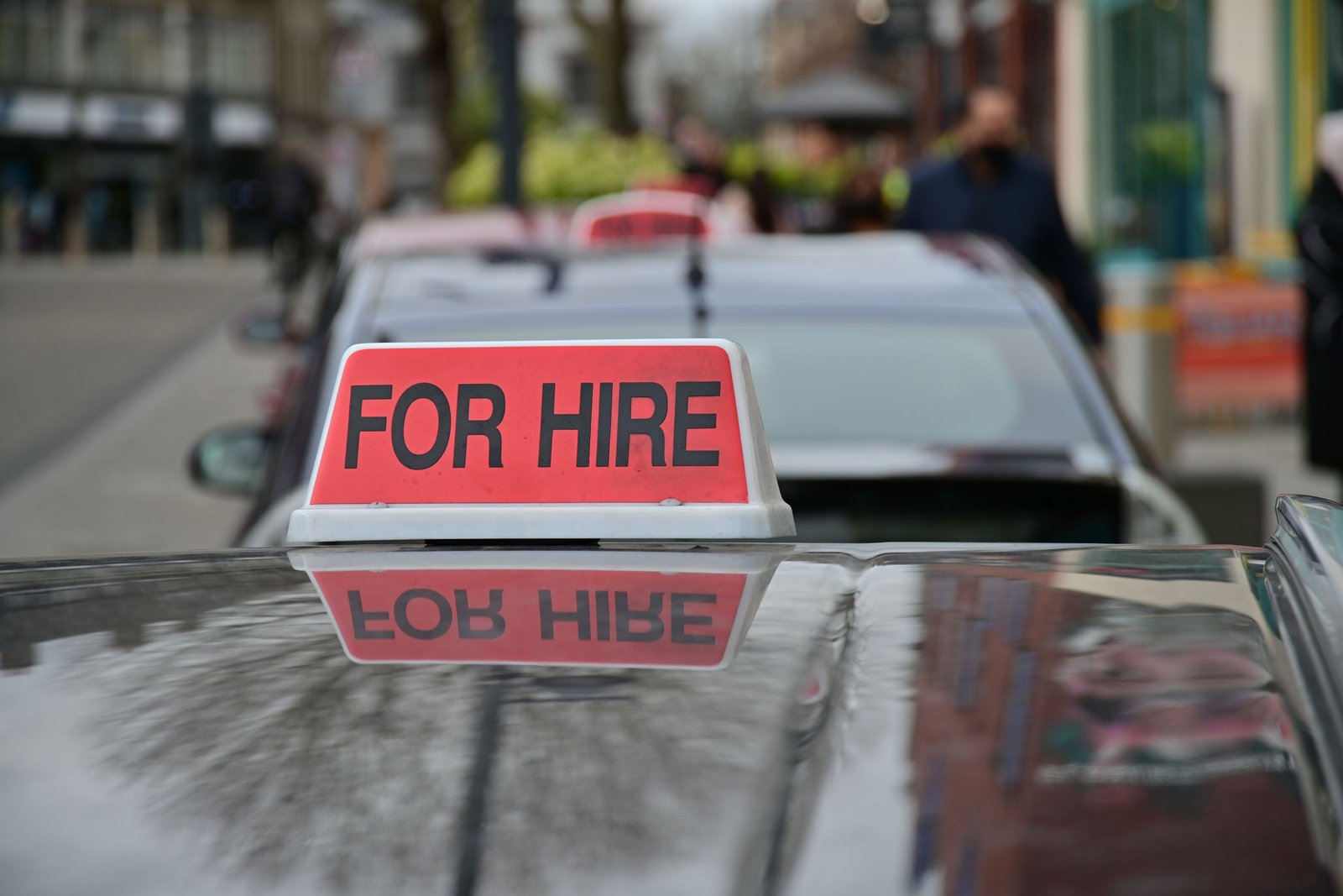 Close-up of a red 'For Hire' sign on a taxi, reflecting the urban street.